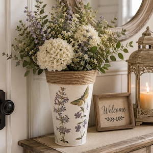 May include: A white tapered metal vase with a jute rope rim, decorated with botanical illustrations of purple flowers and a green moth. The vase is filled with a large arrangement of white hydrangea blossoms, purple lavender-like sprigs, and green foliage.