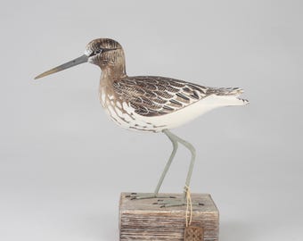 Puede incluir: Escultura de pájaro de madera tallada a mano, con un pico largo y patas delgadas. El pájaro presenta detalles pintados en marrón y blanco, de pie sobre un bloque de madera envejecida. Una pequeña etiqueta cuelga de la base.