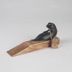 May include: A black seal figurine mounted on a light brown wooden wedge doorstop. A small rectangular wooden tag with the text Archipelago and a geometric pattern is attached to the seal with a thin light-colored string.