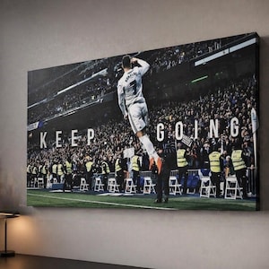 Puede incluir: Impresión en lienzo que muestra a un futbolista con uniforme blanco celebrando un gol. La imagen está ambientada en un estadio lleno de espectadores. Las palabras "KEEP GOING" están impresas en el lienzo.