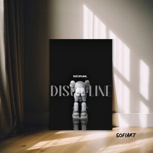 May include: A black canvas art print with a gray cartoon figure covering its face. The word "DISCIPLINE" is printed in large, white letters, with "DISCIPLINE." above it. The artwork is displayed on a wooden floor.