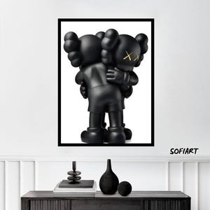 Op de afbeelding: Een ingelijste print met twee zwarte cartoonfiguren die elkaar omarmen. Eén figuur heeft gele 'XX'-ogen. De glanzende figuren steken af tegen een witte achtergrond. De print wordt weergegeven boven een donkerhouten kast met decoratieve items.