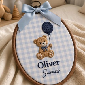 May include: An oval wooden embroidery hoop containing fabric with a light blue and white gingham pattern. The fabric features an embroidered brown teddy bear holding a dark blue balloon, with the name Oliver James stitched in dark blue thread.