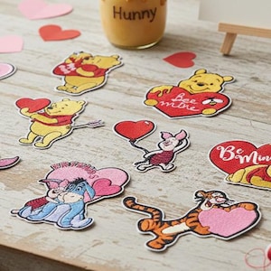 Może przedstawiać: Kolekcja naszywek z motywem Kubusia Puchatka i wycięć w kształcie serca na drewnianej powierzchni. Naszywki przedstawiają postacie takie jak Puchatek, Prosiaczek i Tygrysek, z frazami takimi jak "Bee Mine". Widoczny jest również słoik miodu z napisem "Hunny".