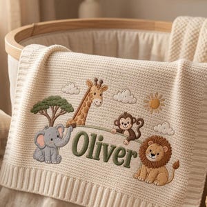 May include: A cream-colored knit baby blanket featuring an embroidered jungle scene with a gray elephant, a giraffe, a brown monkey, and a lion. The name Oliver is stitched in green letters in the center, surrounded by clouds and a sun.