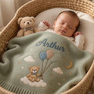 May include: A sage green knitted baby blanket featuring an embroidered teddy bear holding three balloons, white clouds, stars, and a crescent moon. The name Arthur is embroidered in dark blue script above the bear. A small plush teddy bear sits nearby.