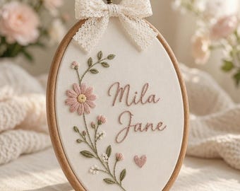 May include: An oval wooden embroidery hoop containing white fabric with the embroidered name "Mila Jane" in pink script. The design features a floral vine with pink daisies, green leaves, and a small pink heart, topped with a cream lace bow.