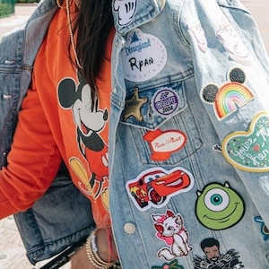 Könnte beinhalten: Hellblaue Jeansjacke mit bunten Disney-Patches, darunter Mickey Mouse, ein Regenbogen und Charakterlogos. Darunter ist ein orangefarbener Pullover mit einem Mickey Mouse-Motiv zu sehen. Auf der Jacke steht "Disneyland Rpan".