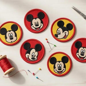 May include: Embroidered Mickey Mouse patches in red and yellow. The patches feature Mickey's face with a black outline and black ears. A spool of red thread, pins, and scissors are also visible.