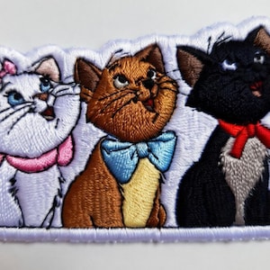 May include: Embroidered patch featuring three cartoon cats. The cats are white, brown, and black, each with unique expressions and accessories. The white cat wears a pink bow, the brown cat a blue bow tie, and the black cat a red bow.