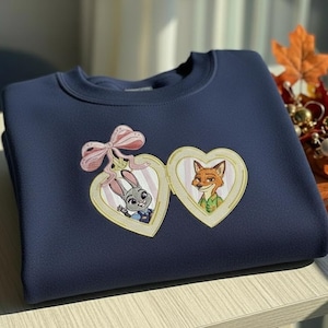 Pu&ograve; includere: Felpa blu navy con un design ricamato di due medaglioni a forma di cuore. Un medaglione mostra un coniglio, l'altro una volpe. Un fiocco rosa &egrave; sopra i medaglioni. La felpa &egrave; su una superficie chiara.