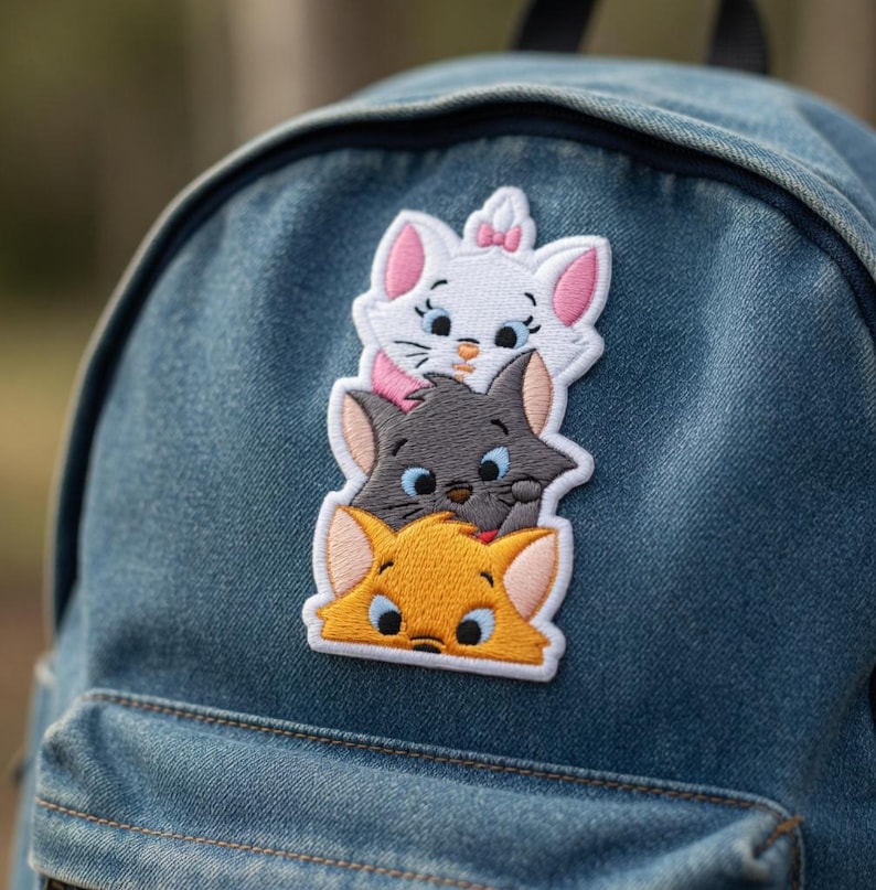 May include: Embroidered patch featuring three cartoon cat faces stacked vertically. The top cat is white with pink accents, the middle cat is gray, and the bottom cat is orange. The patch is attached to a denim backpack.