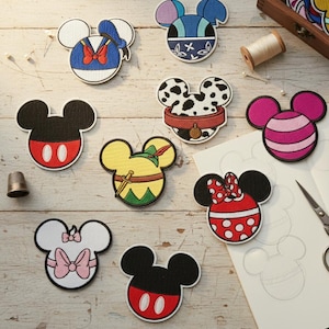 Könnte beinhalten: Eine Sammlung von aufgestickten Aufnähern in Form eines Mäusekopfes, die verschiedene Charakterdesigns zeigen. Die Aufnäher zeigen unter anderem Donald Duck, Mickey Mouse und Minnie Mouse, mit leuchtenden Farben und detaillierten Stickereien. Ein Fingerhut, Faden und eine Schere sind ebenfalls zu sehen.