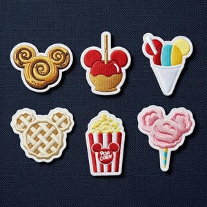 May include: Embroidered patches featuring Mickey Mouse-shaped designs of a cinnamon roll, caramel apple, ice cream cone, waffle, popcorn, and cotton candy. Each patch has a white border and is set against a dark blue background.