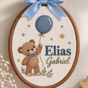 Può includere: Un telaio da ricamo ovale in legno contenente tessuto bianco con un orsacchiotto marrone ricamato che indossa un papillon blu e tiene un palloncino blu. Il testo "Elias Gabriel" è ricamato in blu e verde. Un nastro di raso blu è legato in alto.