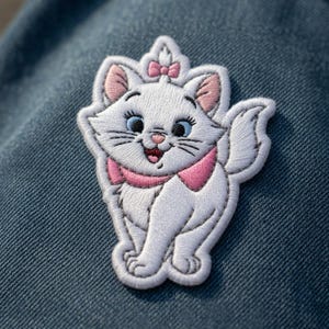 May include: Embroidered patch featuring a white cartoon cat with pink accents on its ears, bow, and neck. The cat has blue eyes and a smiling expression. The patch is on a blue denim background.