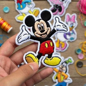 Puede incluir: Parche bordado de Mickey Mouse con detalles en negro, rojo, amarillo y blanco. El parche muestra a Mickey Mouse con los brazos extendidos. Otros parches de personajes de Disney son visibles en el fondo.