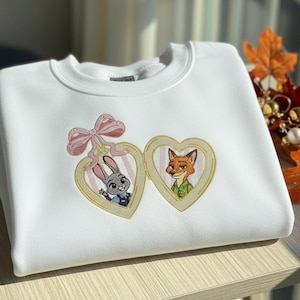 Pu&ograve; includere: Felpa bianca con un disegno ricamato di due personaggi dei cartoni animati in cornici a forma di cuore. Un fiocco rosa &egrave; sopra una cornice. I personaggi sono un coniglio e una volpe. La felpa &egrave; piegata su una superficie di legno.