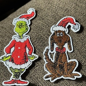 May include: Two embroidered patches featuring Christmas characters. One patch depicts a green character in a red Santa suit and hat. The other patch shows a brown dog wearing a Santa hat and red scarf. Both patches have white borders.