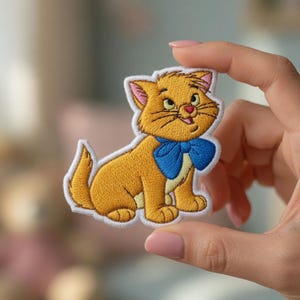 May include: Embroidered patch featuring a cartoon cat with orange fur, a blue bow tie, and a white border. The cat is in a seated position with its tail curled. The patch is held up by a hand.