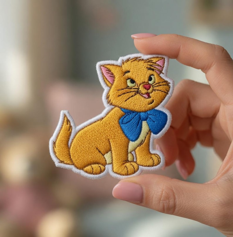 May include: Embroidered patch featuring a cartoon cat with orange fur, a blue bow tie, and a white border. The cat is in a seated position with its tail curled. The patch is held up by a hand.