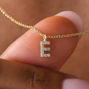 May include: A gold chain necklace featuring a small, rectangular pendant in the shape of the letter E. The pendant is embellished with tiny, clear, sparkling stones set along its front surface, creating a delicate and refined appearance.