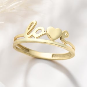 May include: A gold-toned ring featuring a script-style word design that reads love, with the letter v replaced by a small, solid heart shape. The band is thin and polished, showcasing a smooth metallic finish throughout the entire jewellery piece.
