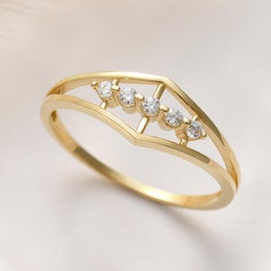 May include: A gold ring featuring a chevron-shaped band with a horizontal bar across the centre. The bar is set with five small, clear, round-cut stones arranged in a row, creating a delicate and symmetrical design on the polished metal band.