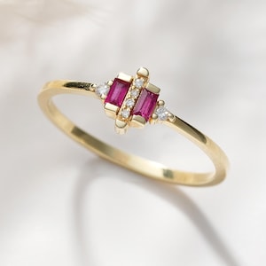 May include: A thin gold ring featuring two vertical baguette-cut red rubies at the center, flanked by small round clear stones and two tiny triangular stones on the outer edges, creating a delicate and symmetrical geometric design on the band.