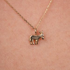 May include: A gold donkey pendant necklace. The donkey charm is detailed with visible legs, ears, and a tail. The necklace chain is delicate and gold, with a small gemstone on the donkey's head.