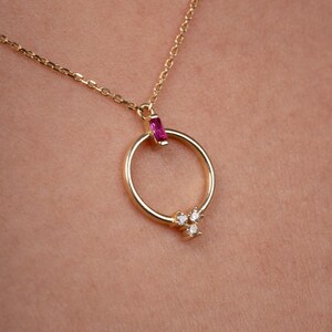 May include: A gold-colored necklace with a circular pendant. The pendant features a small, rectangular pink gemstone at the top and three small white gemstones clustered at the bottom. The necklace chain is delicate and gold-toned.