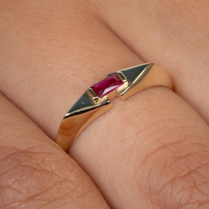 May include: A gold ring with a striking, rectangular red gemstone. The ring showcases a contemporary, geometric design with a split band and angular details. The gemstone is set in a bezel, highlighting its colour and form.