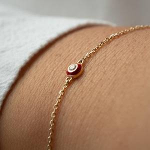 May include: A delicate gold chain bracelet featuring a small, red enamel charm with a central diamond accent. The charm is circular and resembles an eye. The bracelet is shown on a person's arm, with a white shirt in the background.