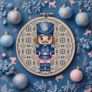 May include: A cross-stitch embroidery of a nutcracker soldier in a wooden hoop. The soldier is dressed in a blue uniform with pink accents. The background is a cream-colored pattern. The image is surrounded by blue Christmas ornaments and decorative elements.