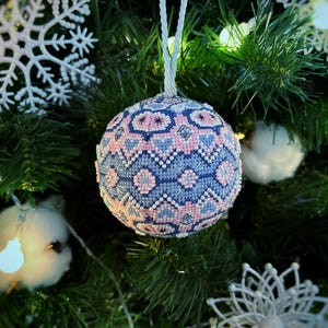 Peut inclure: Une boule de Noël perlée avec un motif géométrique bleu, rose et blanc est suspendue à un sapin de Noël. L'ornement est rond et possède une ficelle blanche pour l'accrocher. L'arrière-plan comprend des aiguilles de pin vertes, des flocons de neige blancs et des guirlandes lumineuses.