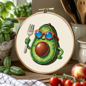 May include: Cross-stitch embroidery in a wooden hoop features a cartoon avocado wearing sunglasses and holding a fork. The avocado is green with a brown pit and water droplets. The background is a light beige fabric.