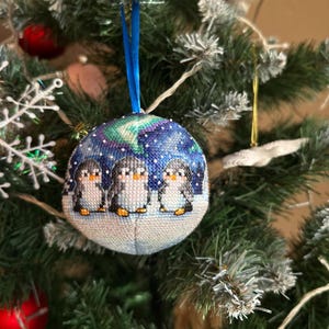 May include: A round Christmas ornament with a blue ribbon. The ornament features a cross-stitch design of three penguins under a starry sky with an aurora borealis effect. The ornament is hanging on a decorated Christmas tree.