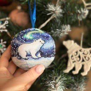 May include: A hand-stitched Christmas bauble depicting a polar bear under a starry sky and aurora borealis. The round bauble has a blue ribbon for hanging. The polar bear is white, and the background is various shades of blue and green.