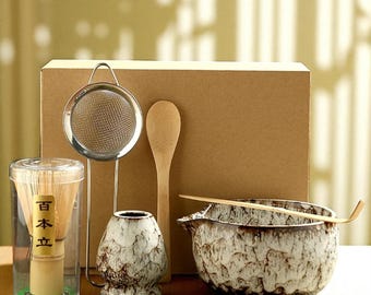 Puede incluir: Un juego de té matcha con un batidor de bambú en un recipiente transparente, un colador de acero inoxidable, un cuenco de cerámica, una cuchara de bambú, una cuchara de madera y un cuenco de té de cerámica. El juego está frente a una caja de cartón.