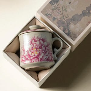 May include: A ceramic mug with a lid, featuring a large pink peony floral design. The mug is presented in a white gift box with a floral pattern. The mug has a curved handle and is approximately 4 inches tall.