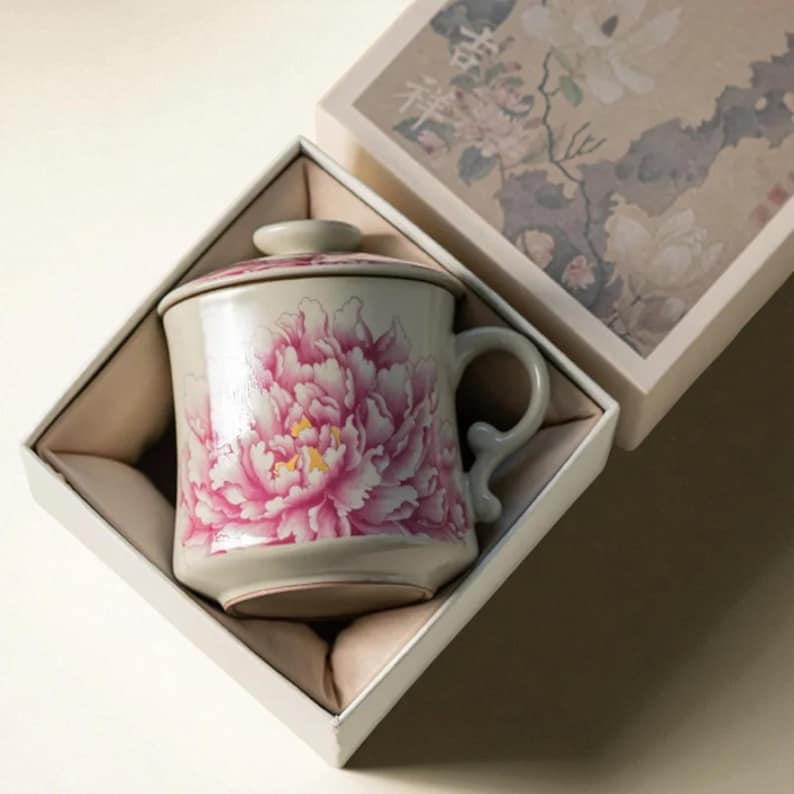 May include: A ceramic mug with a lid, featuring a large pink peony floral design. The mug is presented in a white gift box with a floral pattern. The mug has a curved handle and is approximately 4 inches tall.