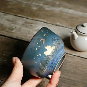 Könnte beinhalten: Eine türkisfarbene Keramik-Teetasse mit einem goldenen Design von Vögeln und Wellen. Die Teetasse hat eine einzigartige Form und wird in der Hand einer Person gehalten. Eine weiße Teekanne mit schwarzem Rand ist im Hintergrund.