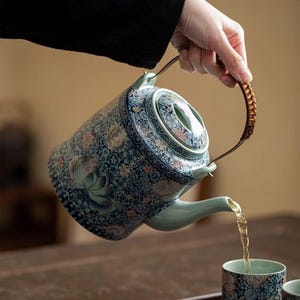 Könnte beinhalten: Eine Keramik-Teekanne mit blau-weißem Blumenmuster, einem hellgrünen Ausguss und einem passenden Deckel. Die Teekanne hat einen Metallgriff mit brauner Umwicklung und gießt Flüssigkeit in eine passende, ebenfalls floral gemusterte Keramiktasse.