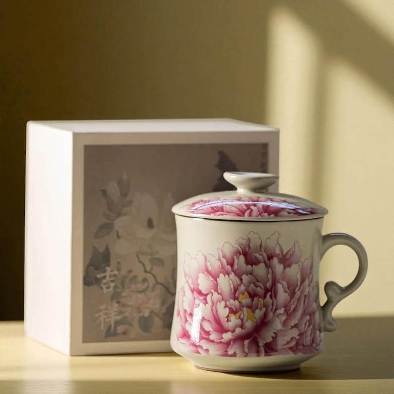 May include: A ceramic mug with a lid, featuring a large pink peony floral design. The mug has a curved handle and is accompanied by a white box with a floral illustration. The mug is likely for tea or coffee.