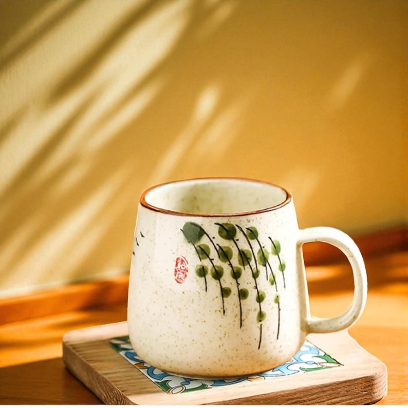 Peut inclure: Mug en c&eacute;ramique cr&egrave;me avec un bord et une anse marron. Le mug pr&eacute;sente un motif peint &agrave; la main de feuilles vertes et un tampon rouge. Il repose sur un dessous de verre en bois avec un insert en carrelage &agrave; motifs. Le mug est pour les boissons chaudes.