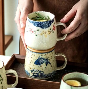 Peut inclure: Deux mugs en c&eacute;ramique, l'un empil&eacute; sur l'autre. Le mug sup&eacute;rieur a une base blanche avec des motifs floraux et bleus. Le mug inf&eacute;rieur a une base beige mouchet&eacute;e avec un motif de poisson bleu et de vagues.