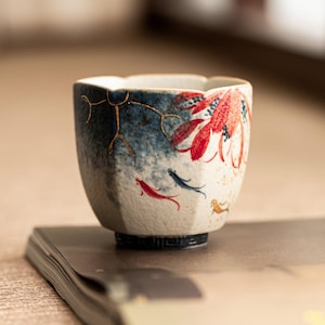 May include: A ceramic tea cup with a textured, off-white base and a dark blue gradient finish. The cup features hand-painted red and blue fish motifs, a large red floral design, and gold-colored line accents. The rim is scalloped and the base is black.