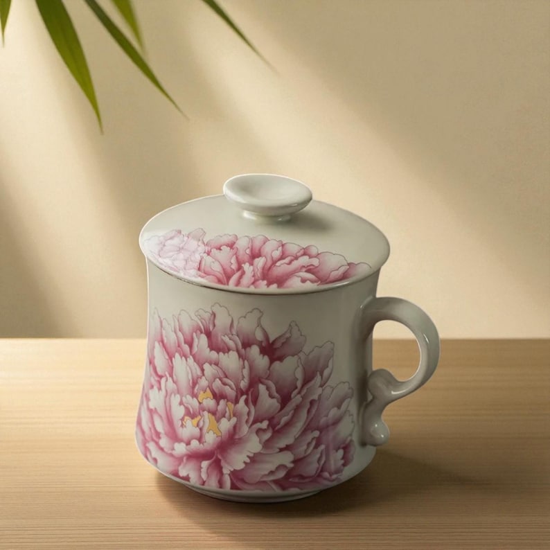 May include: A white ceramic mug with a lid, featuring a large pink peony floral design. The mug has a curved handle and a small knob on the lid. The design is detailed with shades of pink and yellow.