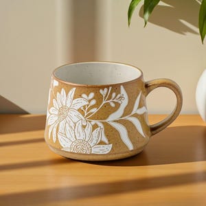 May include: A brown ceramic mug with a white floral design. The mug has a rounded shape, a handle, and a speckled interior. The floral design features large flowers and leaves. The mug is sitting on a wooden surface.