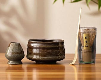 Puede incluir: Un juego de té matcha con un cuenco de cerámica, una pequeña taza de cerámica, un batidor de bambú y una cuchara de bambú. El cuenco y la taza tienen un esmalte marrón y gris. El batidor está en un recipiente transparente.
